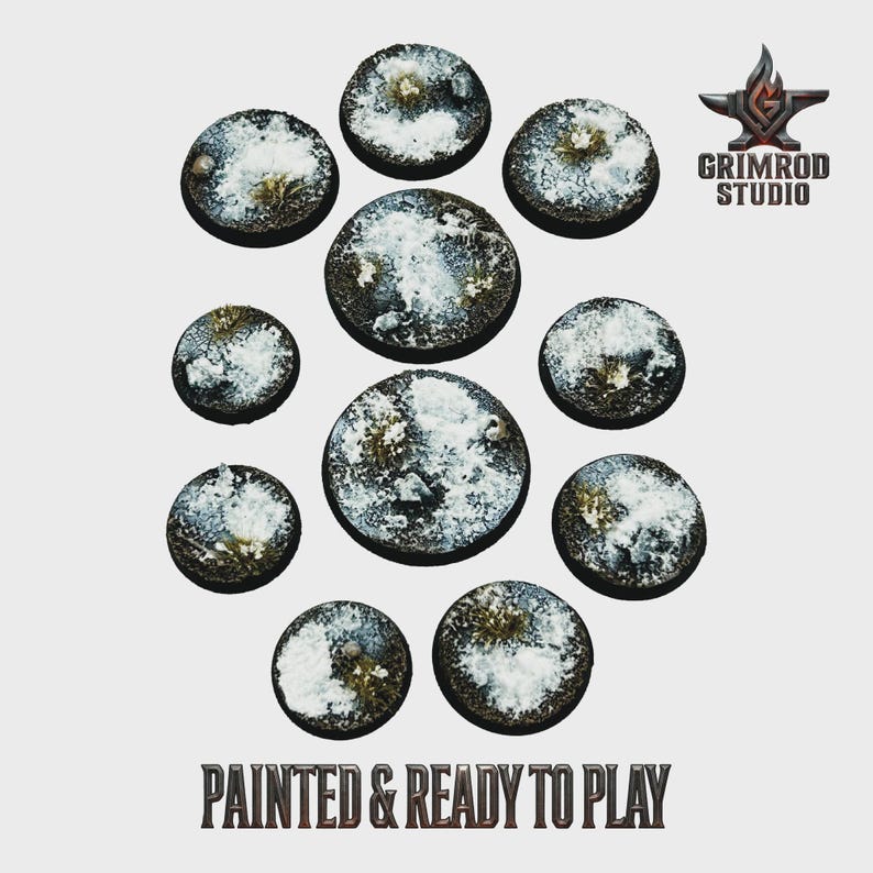 Op de afbeelding: Een verzameling van elf ronde, getextureerde bases voor miniatuurfiguren. De bases zijn beschilderd met een sneeuw- en graseffect, met witte en groene tinten. De tekst "PAINTED & READY TO PLAY" staat onderaan.