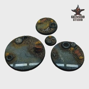 May include: Four circular miniature bases with a post-apocalyptic theme. The bases feature a road surface with white lines, patches of dry grass, and debris like tires and barrels. The Grimrod Studio logo is in the upper right corner.