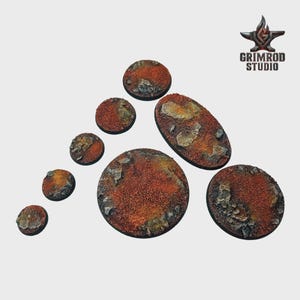 Pu&ograve; includere: Una collezione di basi in miniatura strutturate di varie forme e dimensioni. Le basi presentano una superficie rosso-marrone e granulosa con dettagli simili a rocce incorporati. Il logo Grimrod Studio &egrave; visibile.