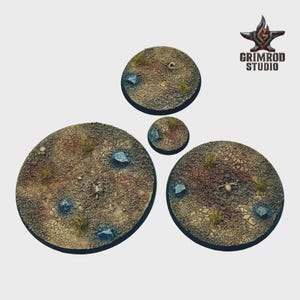 May include: Four circular bases with a desert terrain design. The bases feature a cracked earth texture, small rocks, sparse vegetation, and miniature skeletal remains. The Grimrod Studio logo is in the upper right corner.
