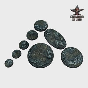 May include: A collection of textured, gray miniature bases in various shapes and sizes. The bases have a rough, rocky appearance, with a mix of dark and light gray tones. The Grimrod Studio logo is in the upper right corner.