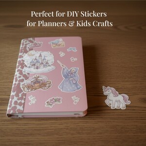 Op de afbeelding: Een roze notitieboek met stickers van een kasteel, koets en fee. Een eenhoornsticker staat naast het notitieboek. Het notitieboek heeft een koeienprint op de randen. De tekst bovenaan luidt "Perfect for DIY Stickers for Planners & Kids Crafts."