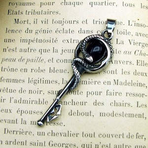 May include: A silver-toned pendant shaped like a key, featuring a coiled snake design around a black gemstone. The pendant is displayed against the backdrop of an open vintage book, creating a gothic or fantasy aesthetic.