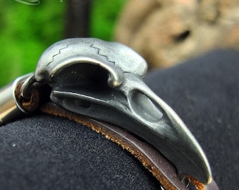 May include: A silver-toned metal bracelet with a detailed bird skull charm. The bracelet is made of brown leather and has a silver buckle.
