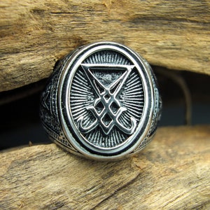 May include: Silver-toned ring with an oval face featuring a detailed, engraved symbol within a sunburst design. The ring has a textured band and is set against a wooden background. The ring's design is intricate and detailed.