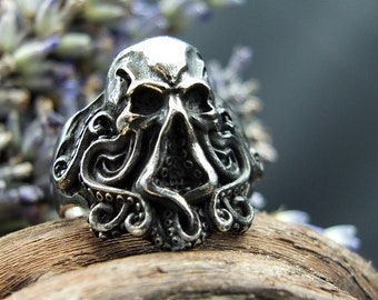 Può includere: Un anello d'argento con un teschio dettagliato e tentacoli, che ricordano un polpo, che si avvolgono attorno al teschio. L'anello è su una superficie di legno.