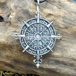 May include: A silver-toned pendant necklace with a compass design featuring Norse runes. The pendant is attached to a black cord.