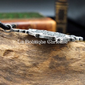 May include: A silver-toned pendant with intricate details, featuring a design of a sword and a shield. The pendant is attached to a black cord.