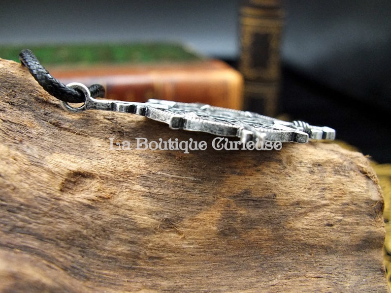 May include: A silver-toned pendant with intricate details, featuring a design of a sword and a shield. The pendant is attached to a black cord.