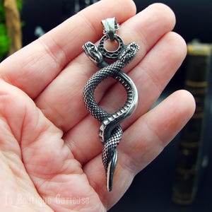 Peut inclure: Pendentif argenté représentant deux serpents entrelacés. Les serpents ont des écailles détaillées et se font face. Le pendentif est conçu pour être porté avec une chaîne. Le pendentif est tenu dans une main.