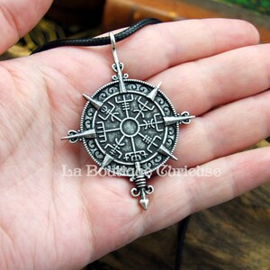 May include: A silver pendant necklace with a compass design featuring a circular compass rose with eight points and a central star. The compass rose is surrounded by a circle with intricate designs and symbols. The pendant is attached to a black cord.