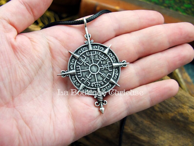 May include: A silver pendant necklace with a compass design featuring a circular compass rose with eight points and a central star. The compass rose is surrounded by a circle with intricate designs and symbols. The pendant is attached to a black cord.
