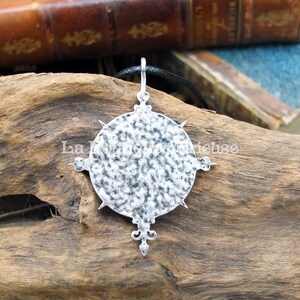 May include: A silver pendant necklace with a circular design featuring a textured surface and a pointed star shape at the bottom. The pendant is hanging from a black cord.