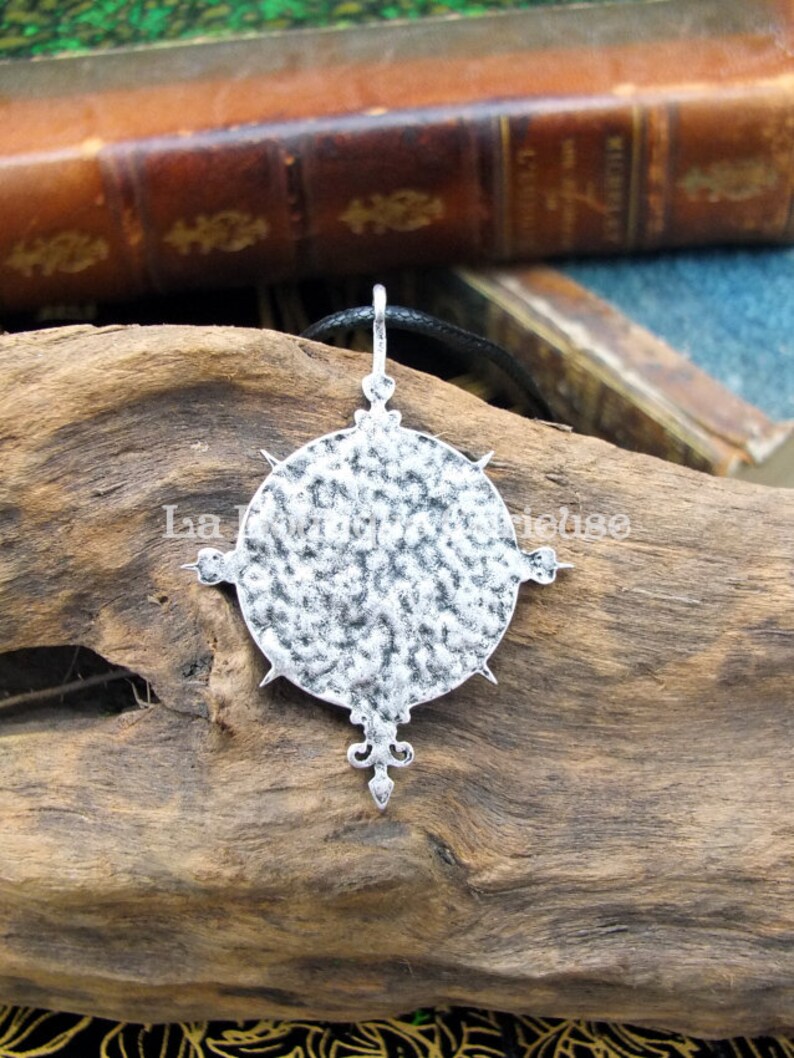 May include: A silver pendant necklace with a circular design featuring a textured surface and a pointed star shape at the bottom. The pendant is hanging from a black cord.