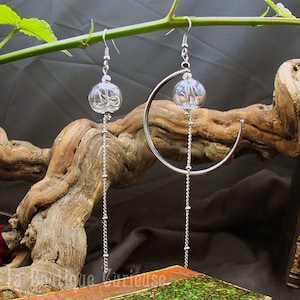 May include: A pair of silver earrings with a crescent moon design and a dangling clear glass orb. The earrings are hanging from a branch.