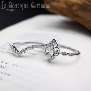 Peut inclure: Deux bagues en argent. L'une présente une pierre précieuse claire en forme de poire, entourée de petites pierres. L'autre est une bande incurvée avec de petites pierres claires. Les bagues sont sur une surface blanche. Le texte "La Boutique Curieuse" est en haut.