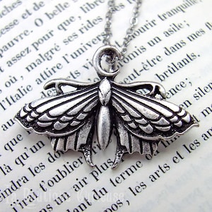 Peut inclure: Un collier avec un pendentif en forme de papillon de nuit argenté. Le pendentif détaillé représente un papillon de nuit aux ailes déployées, suspendu à une fine chaîne. Le collier est posé sur un fond de texte imprimé.