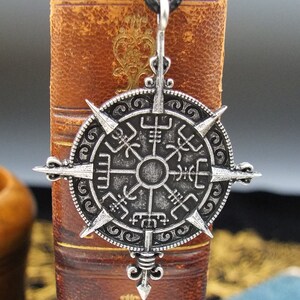 May include: A silver-toned compass pendant with a black cord. The compass features a circular design with eight points and runes around the edge.