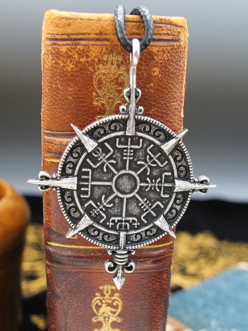 May include: A silver-toned compass pendant with a black cord. The compass features a circular design with eight points and runes around the edge.