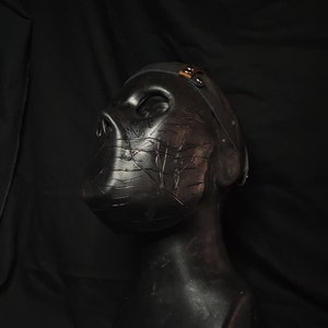 Puede incluir: Una máscara negra con una superficie texturizada y una correa. La máscara está en una cabeza de maniquí negra.