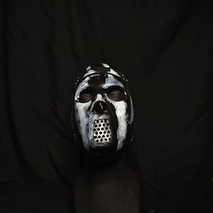 Puede incluir: Una máscara en blanco y negro con una rejilla metálica sobre la boca. La máscara tiene un efecto de pintura agrietada y desgastada y está montada sobre un soporte negro sobre un fondo negro. La máscara tiene aspecto de calavera.