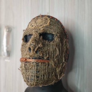 May include: A burlap mask with a copper zipper across the mouth and dark eye sockets. The mask is secured with small pins around the top and is designed to cover the entire face. The mask is a dark brown color.