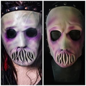 Op de afbeelding: Een wit en paars horrormasker met zwarte details. Het masker heeft een brede mond met een gekarteld, open ontwerp. Het masker is op een zwarte achtergrond.