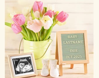 May include: A wooden letter board with a green felt background and white letters spelling "BABY LASTNAME DUE OCT 2025". A framed ultrasound image, a bouquet of pink and white tulips in a yellow bucket, and two white baby booties are on a wooden surface.