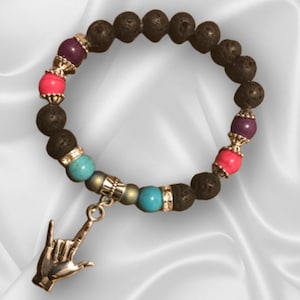 以下が含まれることがあります： ターコイズ、ピンク、パープルのビーズが付いた黒い溶岩岩のブレスレット。ブレスレットには、'愛してる'という手話ジェスチャーをしている手の形のシルバーチャームが付いています。
