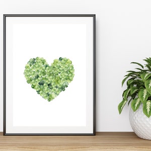 May include: A framed print of a heart shape made of green shamrocks on a white background. The print is hanging on a white wall above a wooden surface with a potted plant.