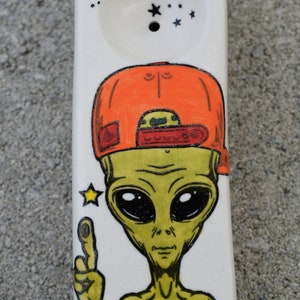 May include: A white ceramic pipe with a green alien wearing an orange baseball cap. The alien has black eyes, a small heart tattoo, and is pointing with one finger. The pipe has a small hole on the top and is decorated with black stars.