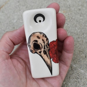 May include: White ceramic pipe with a skull and red flower design. The pipe has a round opening at the top and a small hole on the side.