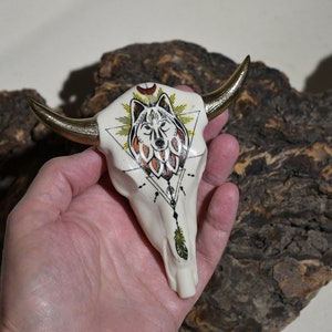 May include: White ceramic bull skull with gold horns, decorated with a black and red wolf head inside a triangle, surrounded by yellow and black sun rays, and a black and white geometric design with a feather hanging from the bottom.
