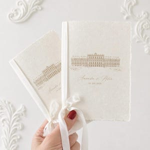 Puede incluir: Dos libretos de papel color crema con bordes irregulares, atados con una cinta blanca. Cada libreto presenta un dibujo lineal en tono dorado de un gran edificio y el texto impreso Amanda & Peter 10. Juli 2026 en fuente script y serif.