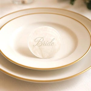 May include: A white plate with a gold rim holds a smaller, round, off-white place card. The place card is inscribed with the word "Bride" in a delicate, cursive font. The plate is set on top of another plate.