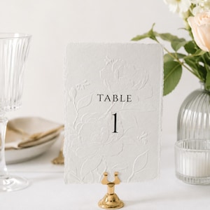 Puede incluir: Una tarjeta rectangular blanca con bordes irregulares que presenta un dise&ntilde;o floral y de abejas en relieve. El texto "TABLE 1" est&aacute; impreso en negro en el centro. La tarjeta se sostiene mediante un peque&ntilde;o soporte de metal dorado pulido.
