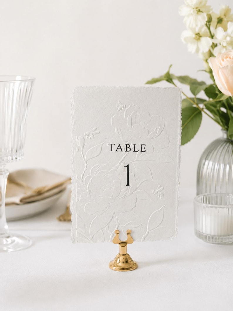 Puede incluir: Una tarjeta rectangular blanca con bordes irregulares que presenta un dise&ntilde;o floral y de abejas en relieve. El texto "TABLE 1" est&aacute; impreso en negro en el centro. La tarjeta se sostiene mediante un peque&ntilde;o soporte de metal dorado pulido.