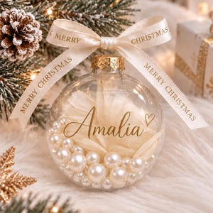 Op de afbeelding: Een doorzichtige glazen kerstbal gevuld met witte kunstbloemblaadjes en diverse witte parels. De naam Amalia staat in goudkleurig schrift op de voorzijde gedrukt. Een crèmekleurig lint met de tekst Merry Christmas is aan de bovenkant gestrikt.