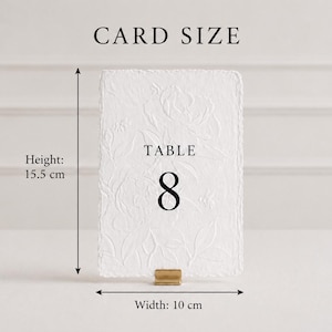 Puede incluir: Una tarjeta de n&uacute;mero de mesa rectangular blanca con un patr&oacute;n floral en relieve texturizado y el texto negro TABLE 8. La tarjeta est&aacute; sujeta por un peque&ntilde;o soporte de metal dorado. Las dimensiones indicadas son 15,5 cm de alto y 10 cm de ancho.