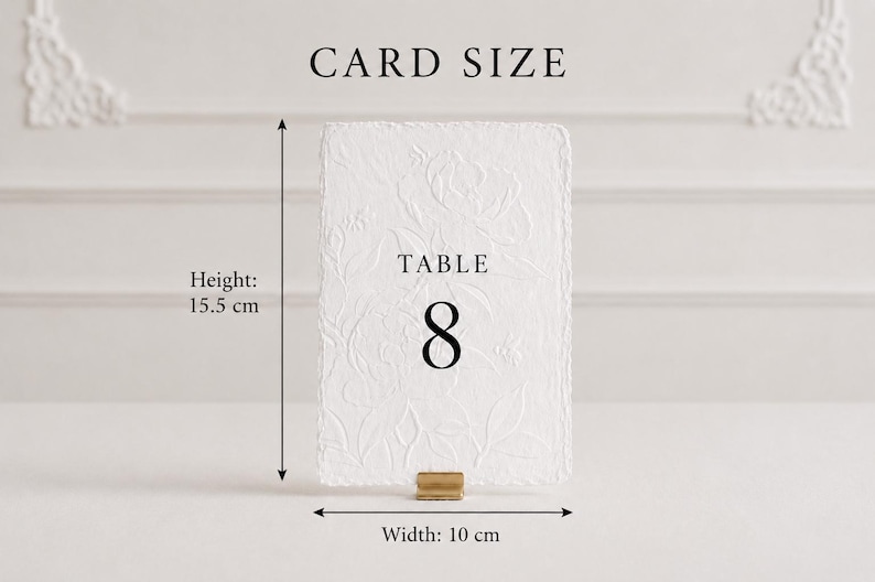 Puede incluir: Una tarjeta de n&uacute;mero de mesa rectangular blanca con un patr&oacute;n floral en relieve texturizado y el texto negro TABLE 8. La tarjeta est&aacute; sujeta por un peque&ntilde;o soporte de metal dorado. Las dimensiones indicadas son 15,5 cm de alto y 10 cm de ancho.