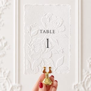 Puede incluir: Una tarjeta rectangular blanca con bordes irregulares que presenta un dise&ntilde;o floral y de abejas en relieve seco. El texto "TABLE 1" est&aacute; impreso en negro en el centro. La tarjeta est&aacute; sujeta por un peque&ntilde;o soporte de metal dorado brillante.