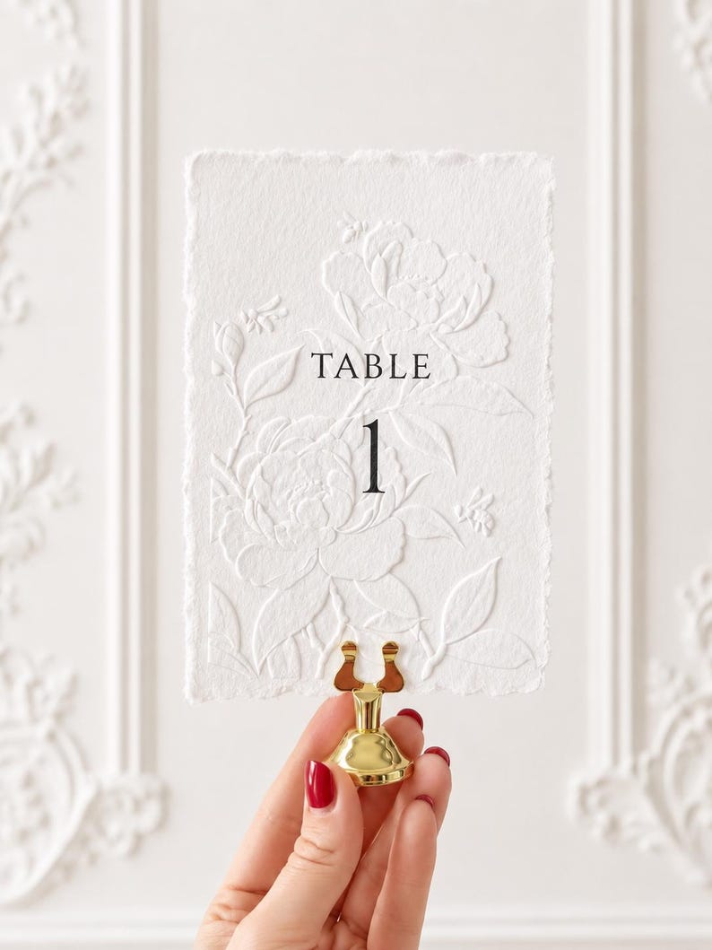 Puede incluir: Una tarjeta rectangular blanca con bordes irregulares que presenta un dise&ntilde;o floral y de abejas en relieve seco. El texto "TABLE 1" est&aacute; impreso en negro en el centro. La tarjeta est&aacute; sujeta por un peque&ntilde;o soporte de metal dorado brillante.
