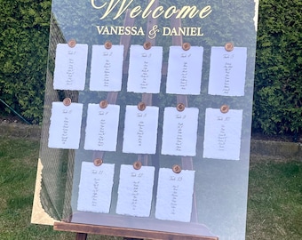 May include: A clear acrylic wedding seating chart with gold lettering. The chart is on an easel and has the names Vanessa and Daniel at the top. The chart lists tables 1 through 13 with guest names on each table.