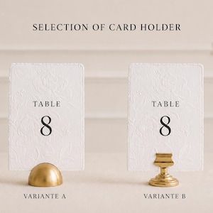 Puede incluir: Dos tarjetas rectangulares blancas de n&uacute;mero de mesa con patrones florales en relieve y el texto TABLE 8 en negro. La tarjeta de la izquierda se sostiene con un soporte dorado en forma de c&uacute;pula etiquetado VARIANTE A, y la de la derecha con un soporte de pedestal dorado VARIANTE B.