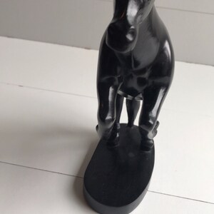 Puede incluir: Una figura de caballo negro con un acabado brillante. La escultura est&aacute; sobre una base ovalada negra. El caballo est&aacute; posando con las patas delanteras levantadas, dando la impresi&oacute;n de movimiento. El fondo es una superficie blanca y una pared blanca.