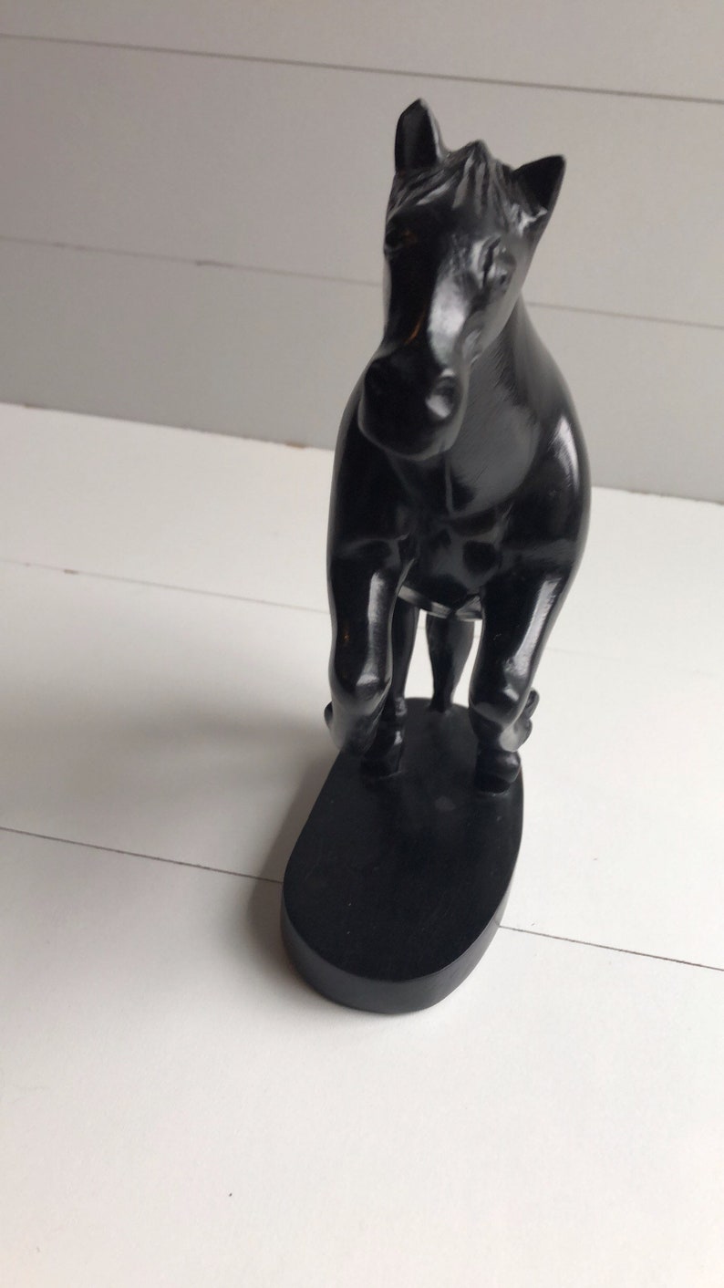 Puede incluir: Una figura de caballo negro con un acabado brillante. La escultura est&aacute; sobre una base ovalada negra. El caballo est&aacute; posando con las patas delanteras levantadas, dando la impresi&oacute;n de movimiento. El fondo es una superficie blanca y una pared blanca.