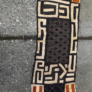 Puede incluir: Un textil largo y estrecho con un patrón geométrico en tonos marrones, naranjas y blancos. El textil está hecho de tejido tejido y tiene una textura rugosa.