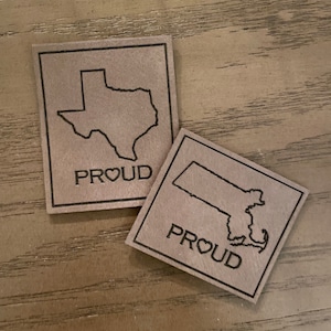 May include: Two brown leather patches with black outlines. The patches feature the outlines of the states of Texas and Massachusetts. Each patch has the word "PROUD" with a heart symbol in the middle of the word.