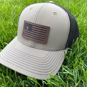 May include: A khaki-colored trucker hat with a black mesh back. The front features a brown leather patch with a deer head silhouette and a flag design. The hat is sitting on a bed of green grass.