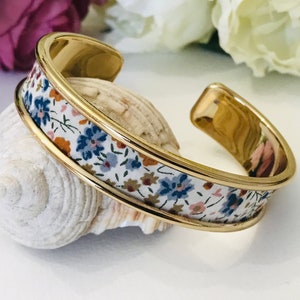Könnte beinhalten: Ein goldenes Armreif-Armband mit einem floralen Stoffband. Der Stoff ist weiß mit blauen, rosa und braunen Blumen.